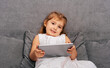 © dvulikaia - Little girl sitting on sofa using modern gadget to watch videos and play games.