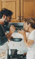 One happy real couple at home enjoying life together and relationship. Man giving food to woman to taste. Male people cooking for his wife. Male and female enjoying leisure activity indoor in kitchen