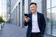 © Tetiana - Smiling young asian business man lawyer standing outside office center and using mobile phone, hand in pocket