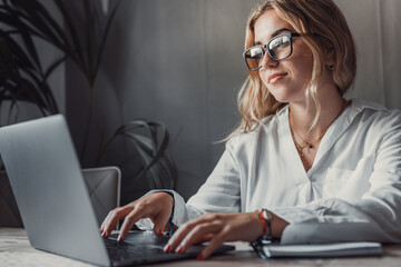 Happy young blonde business woman entrepreneur using computer looking at screen working in internet sit at office desk, smiling millennial female professional employee typing email on laptop at work