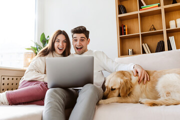 Young overjoyed couple using laptop computer shopping online sitting on comfortable sofa at home. Excited man and woman receive payment, win money 