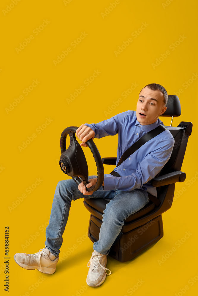 Young man with steering wheel in car seat on yellow background