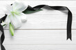 Frame made of beautiful lily flowers and black funeral ribbon on white wooden background