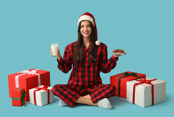 Poster - Beautiful young woman in checkered pajamas with sweet cookies, glass of milk and Christmas gift boxes on blue background