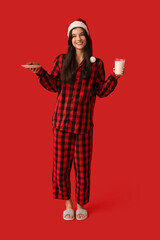 Poster - Beautiful young woman in checkered pajamas with sweet cookies and glass of milk on red background