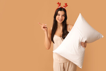 Poster - Beautiful young woman in silky pajamas and reindeer horns with soft pillow pointing at something on brown background