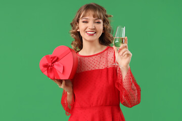 Poster - Beautiful young woman with glass of champagne and gift box on green background. Valentine's Day celebration