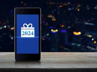 Gift box happy new year 2024 flat icon on modern smart mobile phone screen on wooden table over blur colorful night light city tower and skyscraper, Business happy new year 2024 shop online concept