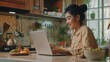 Smiling beautiful young woman using laptop while having breakfast at home kitchen