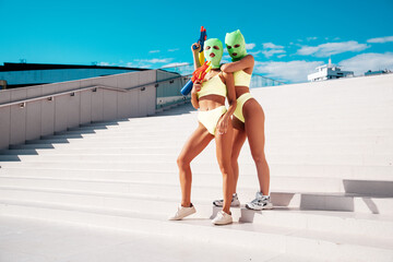 Two beautiful sexy women in green underwear. Models wearing bandit balaclava mask. Hot seductive female in nice lingerie posing in the street at sunny day, blue sky. Crime and violence. Hold water gun