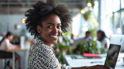 Canvas Print - Smiling young African American businesswoman working on a laptop at her desk in a bright modern office with colleagues in the background