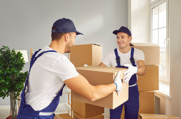 Team of two young happy cheerful professional workers of moving service in overalls carrying and delivering cardboard boxes in new apartment. Move, moving day and relocation concept.