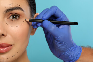 Wall Mural - Doctor with pencil preparing patient for cosmetic surgery operation on light blue background, closeup