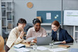 © Seventyfour - Medium shot of three cheerful diverse business women sitting at office desk looking at laptop