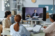 © Seventyfour - Medium shot of company employees at video conference call, focus on big plasma screen with smiling red-haired business lady holding papers and talking