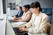 © Seventyfour - Side view of brunette business woman in glasses wearing beige suit working on laptop while sitting at table in company office with colleagues in background