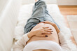 Cropped color photo of pregnant woman in unbuttoned blue jeans laying on white couch with hands on her bare pregnant belly aesthetic