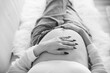 Cropped black and white photo taken from above of pregnant woman in unbuttoned blue jeans laying on white couch with hands on her bare pregnant belly aesthetic
