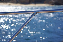 Macro Of Shiny Metal Rail On Boat, Ocean And Rocky Beach In Background With Bokeh
