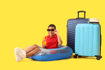 Poster - Happy African-American boy sitting on inflatable ring with cocktail and suitcases against yellow background