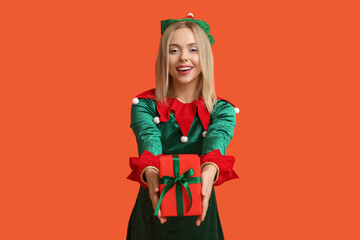 Wall Mural - Young woman in elf costume with Christmas gift box on orange background