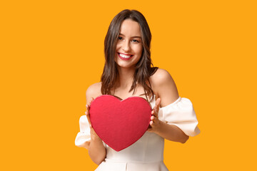 Canvas Print - Beautiful young woman with gift box in shape of heart on yellow background. Valentine's Day celebration