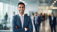 Portrait Of A Handsome Smiling Asian Indian Businessman Boss In A Suit Standing In His Modern Business Company Office. His Workers Standing In The Blurry Background
