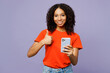 Little kid teen girl of African American ethnicity wear orange t-shirt hold in hand use mobile cell phone show thumb up isolated on plain pastel light purple background. Childhood lifestyle concept.