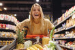 © ViDi Studio - Young excited cheerful customer woman wearing casual clothes shopping at supermaket store grocery shop buying with trolley cart choose products inside hypermarket. Purchasing food gastronomy concept.
