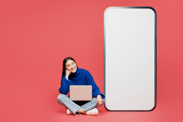 Wall Mural - Full body young IT woman of Asian ethnicity wear blue sweater casual clothes sit use laptop pc computer big huge blank screen mobile cell phone smartphone with area isolated on plain pink background.