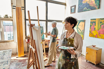 Wall Mural - thoughtful mature woman in apron looking at easel near female friend in modern spacious art workshop