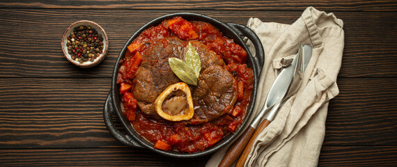 Wall Mural - Traditional Italian dish Ossobuco all Milanese made with cut veal shank meat with vegetable tomato sauce served in black casserole pan top view on rustic brown wooden background.