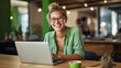 © MP Studio - Cheerful professional woman wearing glasses and a green blouse is sitting at a desk with a laptop and holding coffee cup