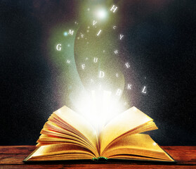 Open book with magic light and glowing letters flying out of it on wooden table against black background