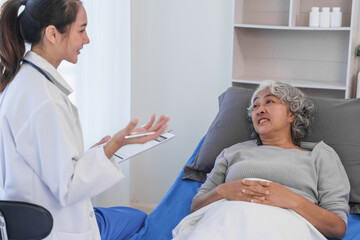 Wall Mural - Elderly Asian woman lying in hospital bed looking unwell, with young Asian doctor take a history of illness Health check and encouragement.