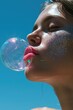 © Glittering Humanity - Close-up of a woman blowing a bubble, featuring sparkling glitter makeup against a blue sky