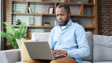 Confident mature african american man typing on laptop sitting on sofa in living room at home office. Thoughtful freelancer works on a computer remotely. A businessman is busy with a startup project