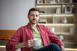 Positive carefree middle-aged bearded man resting on chair at apartment. Cheerful male sitting at home, holding cup, drinking tea, looking at camera friendly smiling. Wellbeing, slow living concept