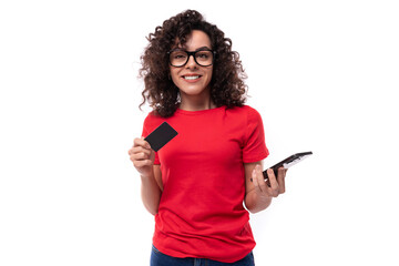 Wall Mural - young pretty curly brunette woman in red t-shirt holding credit card mockup and smartphone