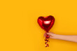 Hand hold red inflatable foil balloon in a heart shape. Festive concept in front of yellow background.