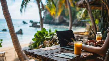 Digital Nomad Working in a Tropical Location - person using a laptop at a wooden table on a tropical beach, with a notebook and a glass of orange juice