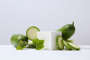 Wall Mural - A white cube placed on white surface with green leaves and winter melon slices. Glamour minimal pedestal for beauty, cosmetic product presentation