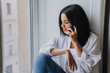 © Iona - Serene woman in white shirt speaking on smartphone by the window, in a relaxed pose.