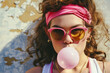 © J S - 1980s retro teenage girl wearing headband with colorful sunglasses and blowing chewing gum bubble, grunge plaster wall in background