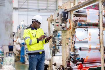 Wall Mural - African male engineer department head Pressing laptop to check machine system and gear circuit board and making bags. Wearing a vest and safety helmet in a plastic and steel industry.