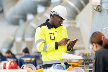 Canvas Print - Close-up shot in a plastic material production factory. African male engineer is looking after a laptop and working near several employees. Wearing a vest, safety helmet. Around are working machines.