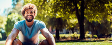 A Lifelike Curly-haired Young Man In A Blue T-shirt Stretches Out On A Yoga Mat In A Sunny Park, With Trees In Soft Focus In The Background.Close-up Shot.Fitness Exercise, Active And Healthy Lifestyle