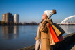 Happy woman in warm clothing with shopping bags enjoys standing  by river on sunny winter day. 