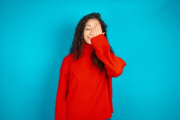 Beautiful teen girl wearing knitted red sweater over blue background covering one eye with her hand, confident smile on face and surprise emotion.