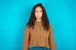Dissatisfied Beautiful teen girl wearing knitted sweater over blue background purses lips and has unhappy expression looks away stands offended. Depressed frustrated model.
